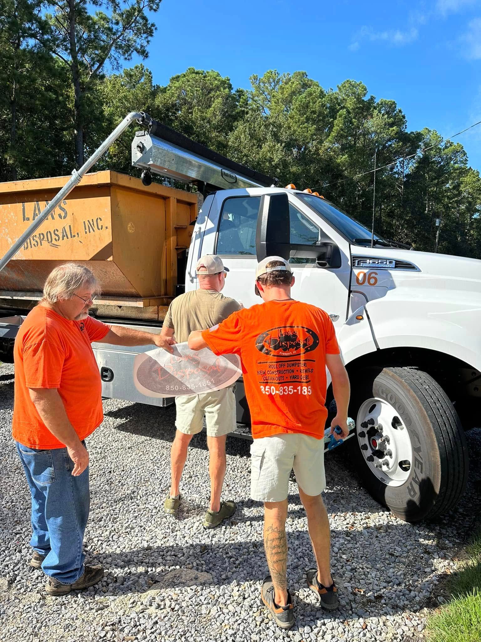 Three people in orange shirts and a tan hat stand by a white truck on gravel, reading a map near a dumpster.