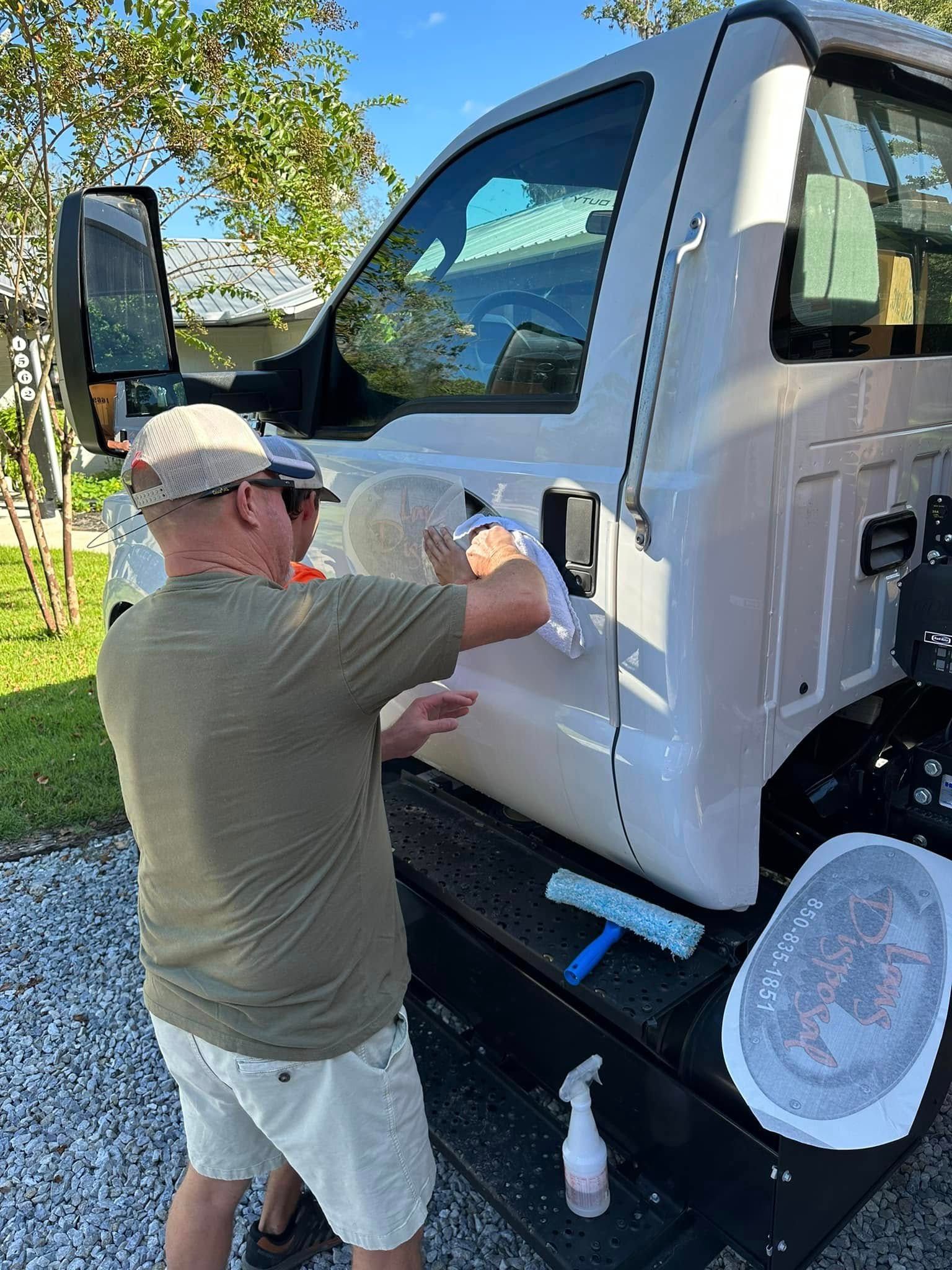 Man washing a white semi-truck cab outdoors with a spray bottle and cloth.