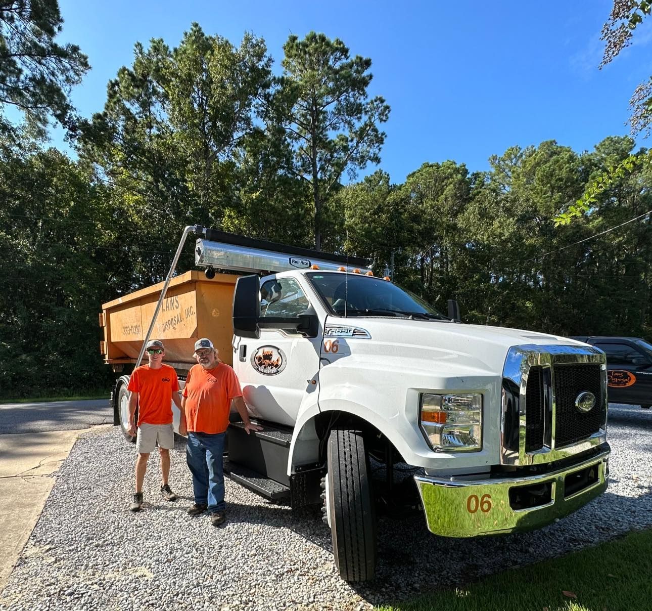 Two people in orange shirts standing beside a white dump truck on a sunny gravel road, surrounded by trees
