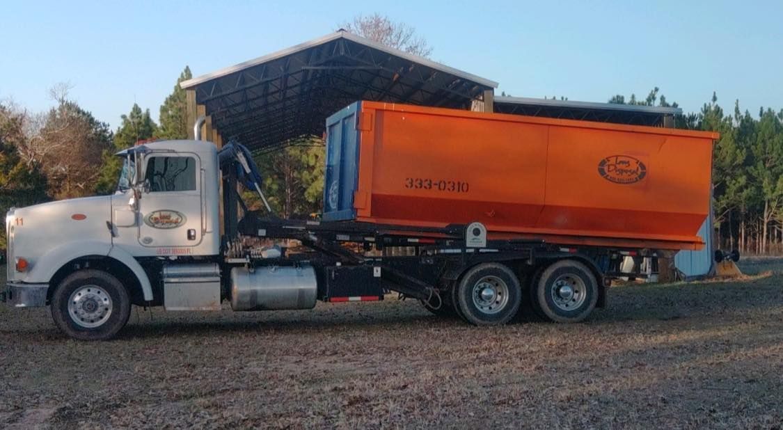 White dump truck with orange bed parked on gravel outdoors near trees and a shed