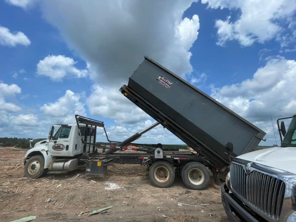 Dump truck unloading gravel at a construction site, with a white truck nearby under a cloudy sky