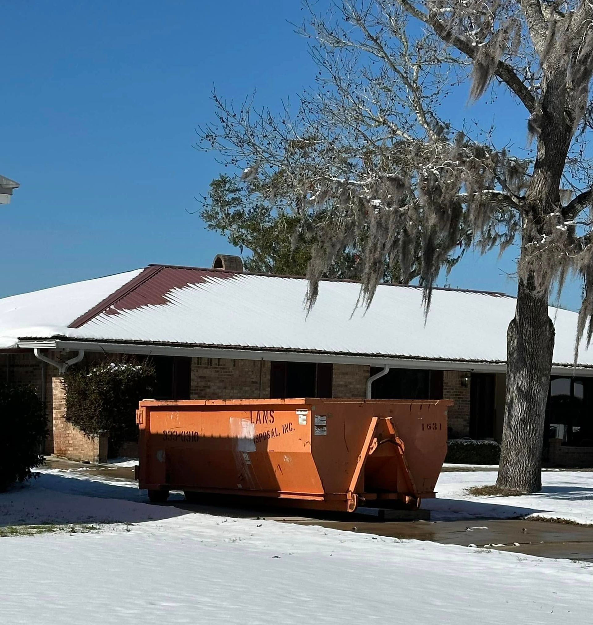 Snowy driveway with an orange dumpster in front of a house with a white roof and bare trees Snowy driveway with an orange dumpster in front of a house with a white roof and bare trees