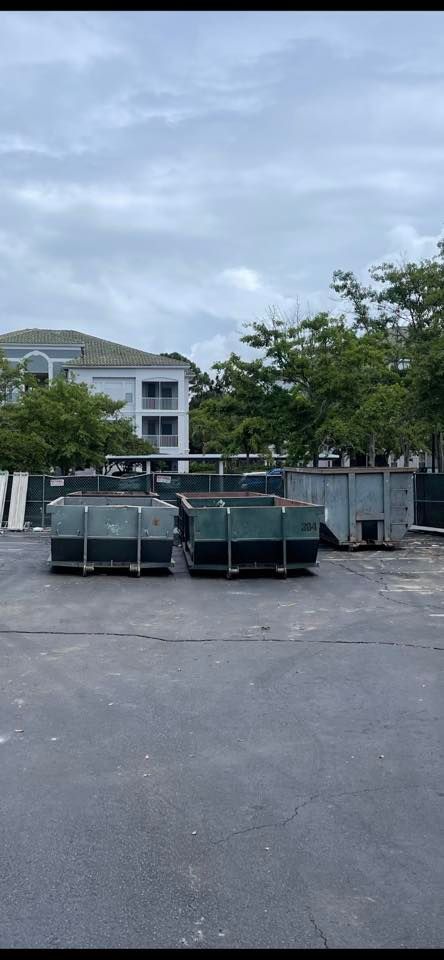 Row of green dumpsters in a parking lot with a building and trees in the background