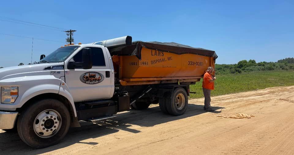 White dump truck with orange bed on a dirt road, with a worker standing beside it under a sunny sky