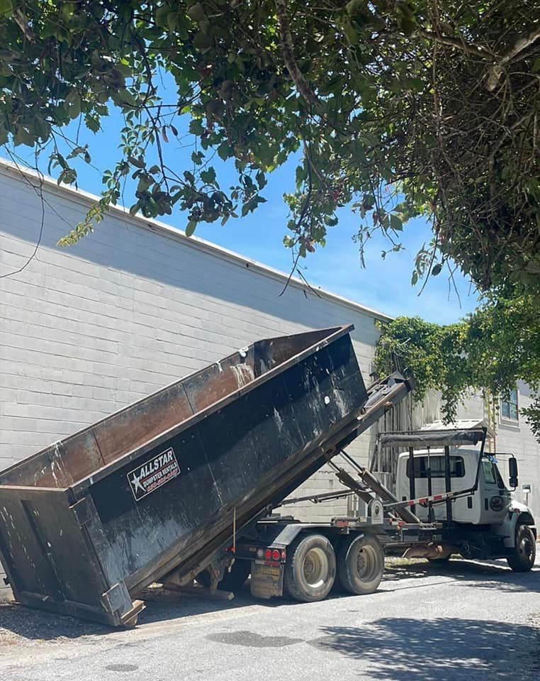Dump truck tipping black dumpster beside a white building under trees on a sunny day