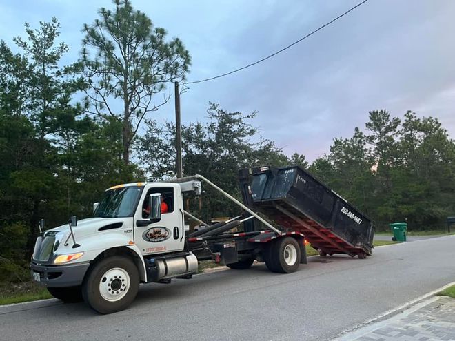 White tow truck hauling a black dumpster trailer on a roadside near trees White tow truck hauling a black dumpster trailer on a roadside near trees