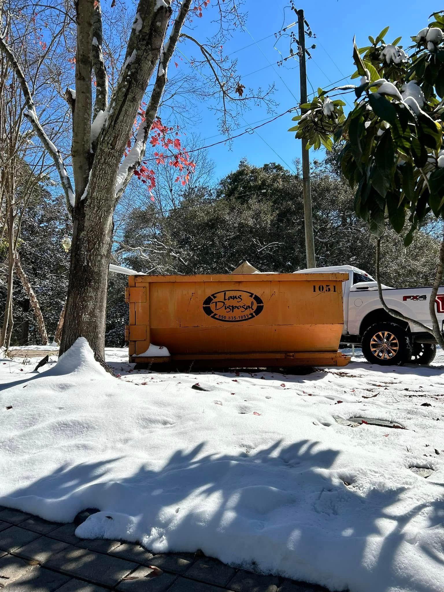 Orange dumpster in snowy yard beside trees and a white pickup truck under a blue sky
