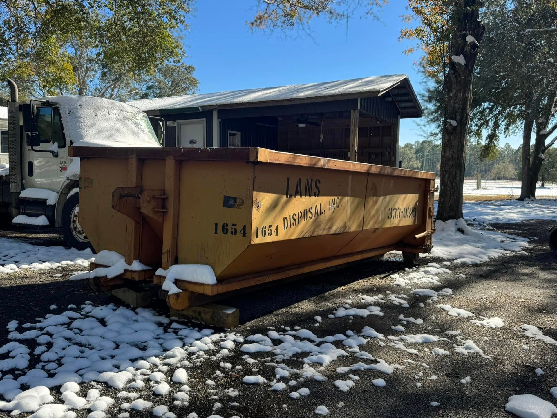 Large orange dumpster beside a truck in a snowy yard under trees Large orange dumpster beside a truck in a snowy yard under trees