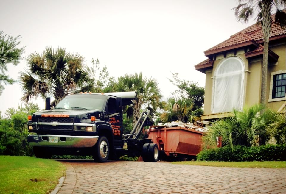 Tow truck hauling a dumpster beside a house with palm trees and a tiled driveway