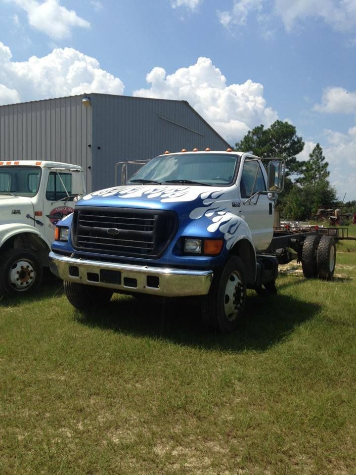 Blue tow truck parked on grass beside a gray warehouse under a partly cloudy sky.