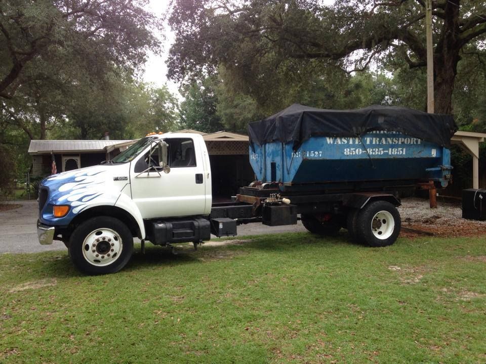 White dump truck with blue cargo bed parked on grass near trees and a house
