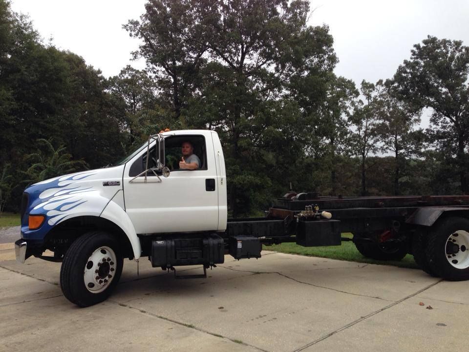 White truck chassis on a driveway with a driver in the cab, trees in the background.