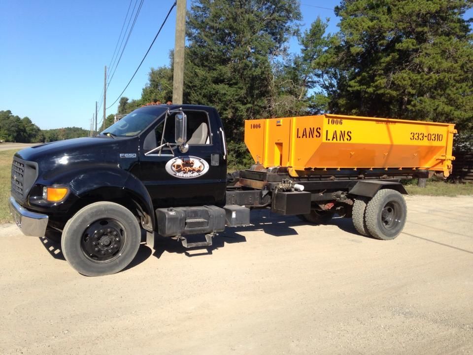Black dump truck with orange Lams container parked on a sunny roadside near trees