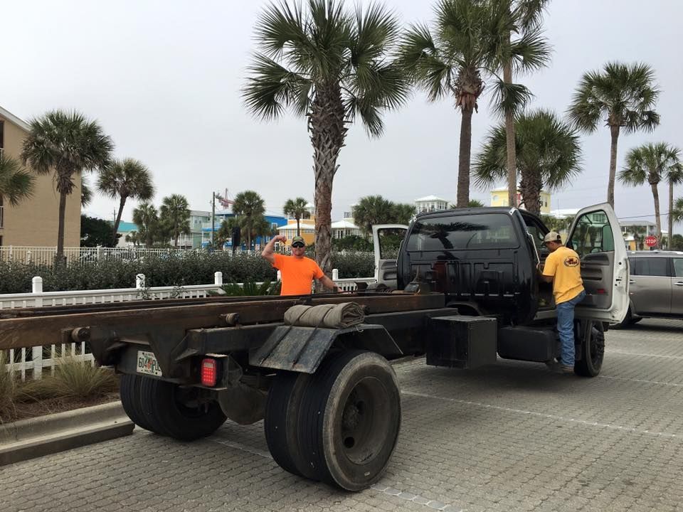People unloading a black truck on a palm-lined street beside a parked van