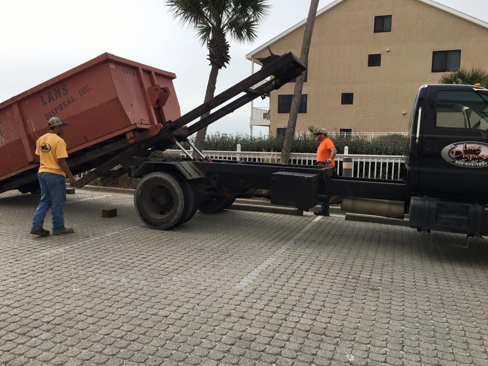 Tow truck lifting a red dumpster in a paved lot, with two workers nearby and a building in the background Tow truck lifting a red dumpster in a paved lot, with two workers nearby and a building in the background