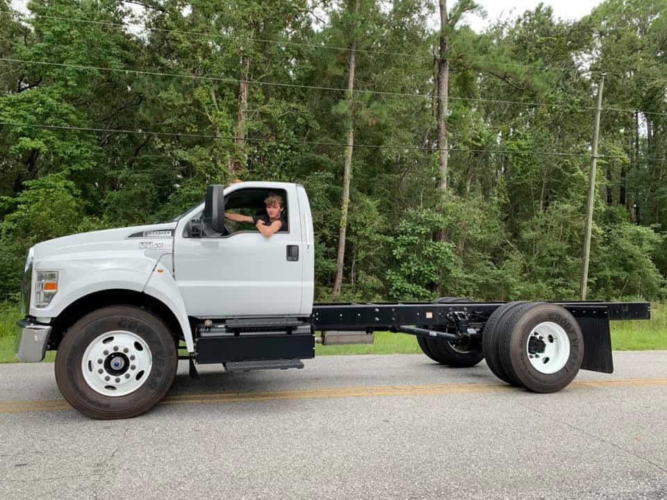 White cab-over flatbed truck parked on a road, driver visible, with trees in the background.