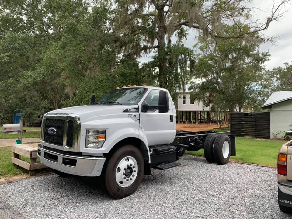 White Ford cab-over truck chassis parked on gravel beside trees and a house