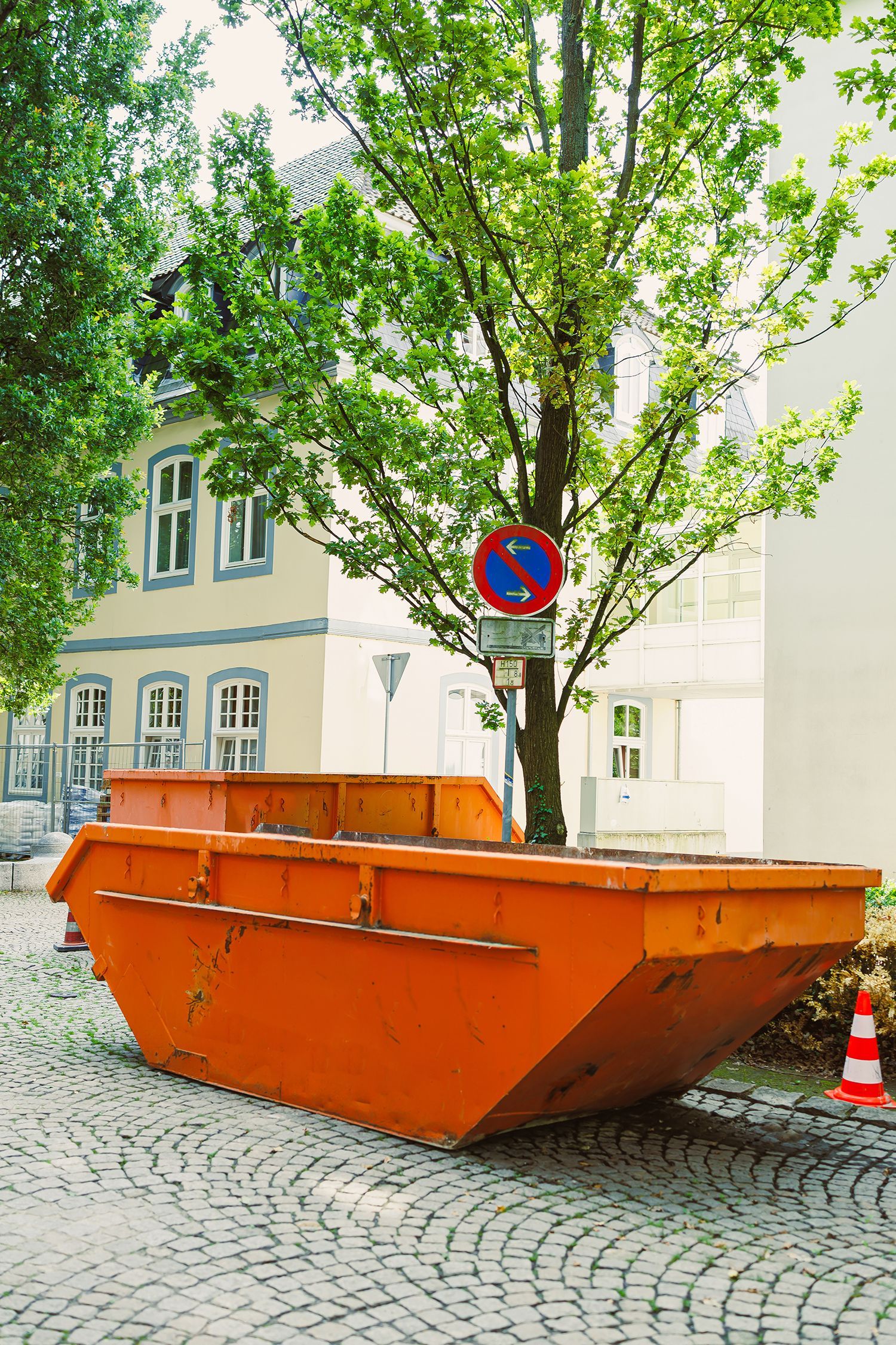Orange dumpster on cobblestone street beside a tree and no-parking sign, with buildings in the background Orange dumpster on cobblestone street beside a tree and no-parking sign, with buildings in the background
