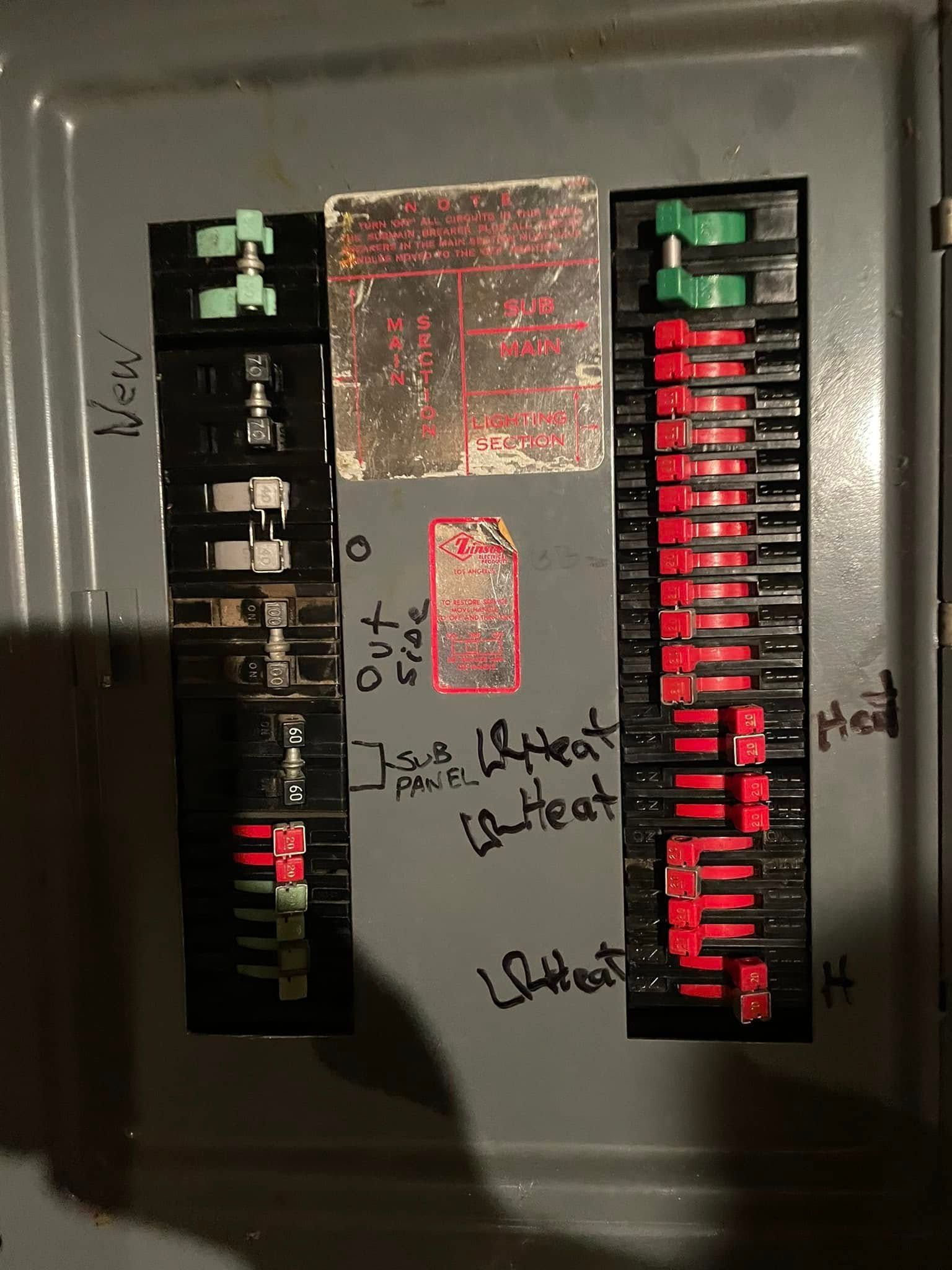 Circuit breaker panel with labeled switches, mostly red and green toggles, and handwritten notes on the center cover.