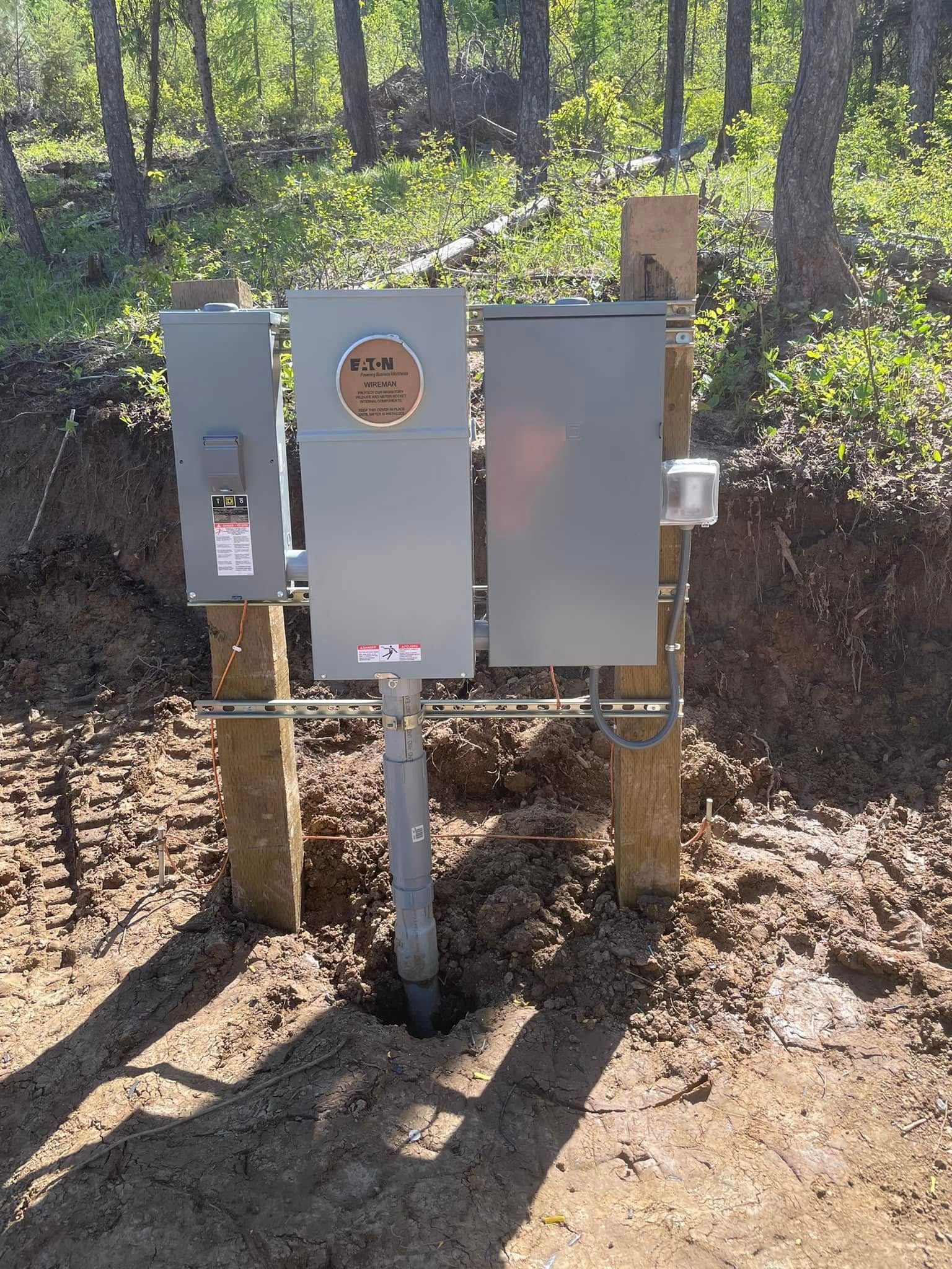 Gray utility boxes on a post in a wooded outdoor area with dirt ground and trees.