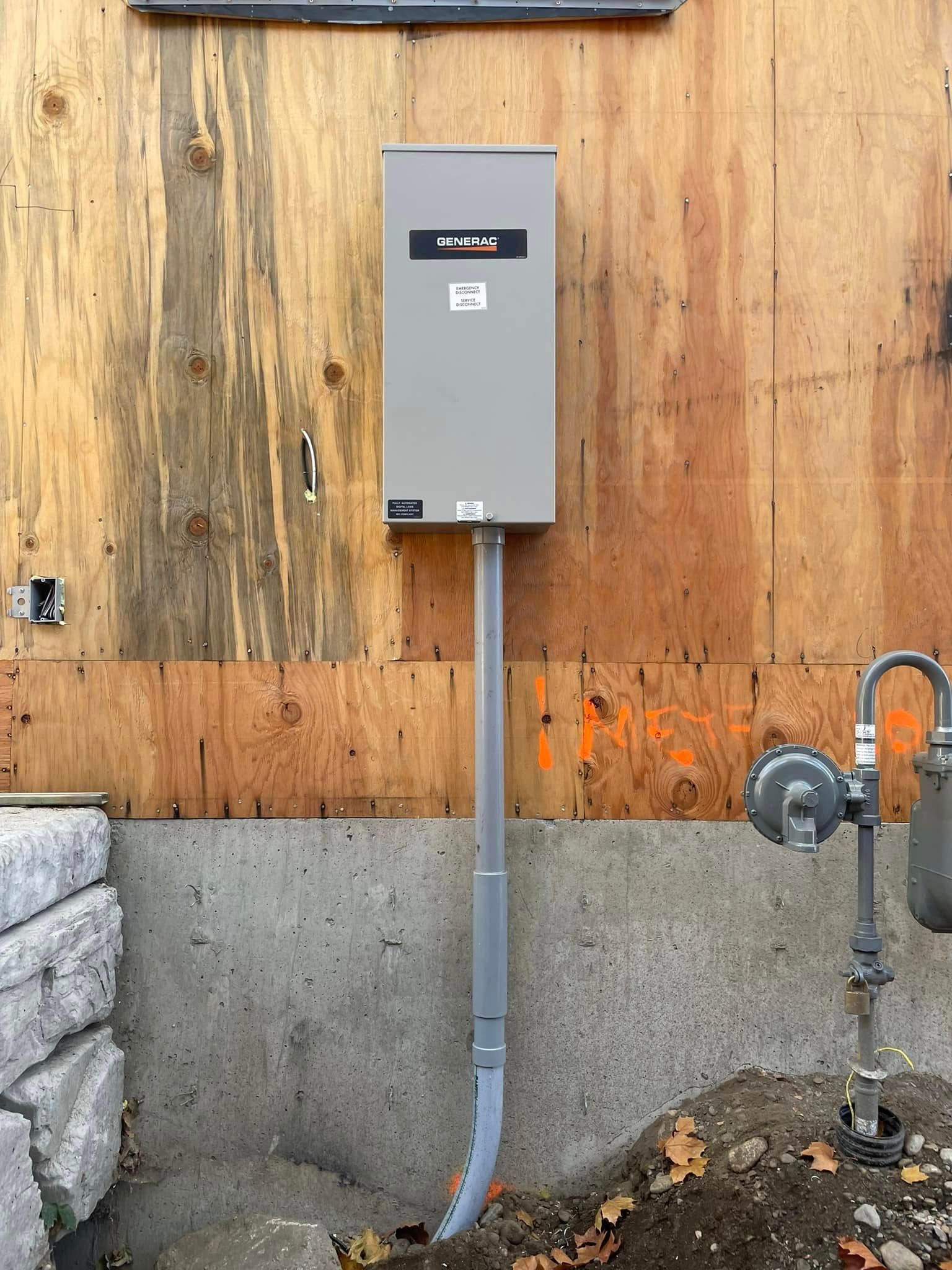 Gray electrical panel mounted on plywood wall above concrete foundation, with conduit running down to the ground