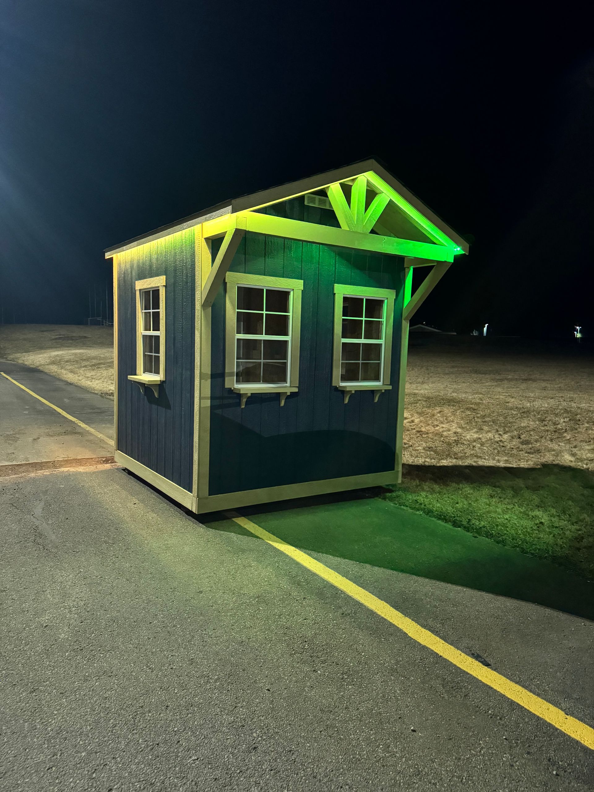 Small blue kiosk with neon green roof lights beside a road at night