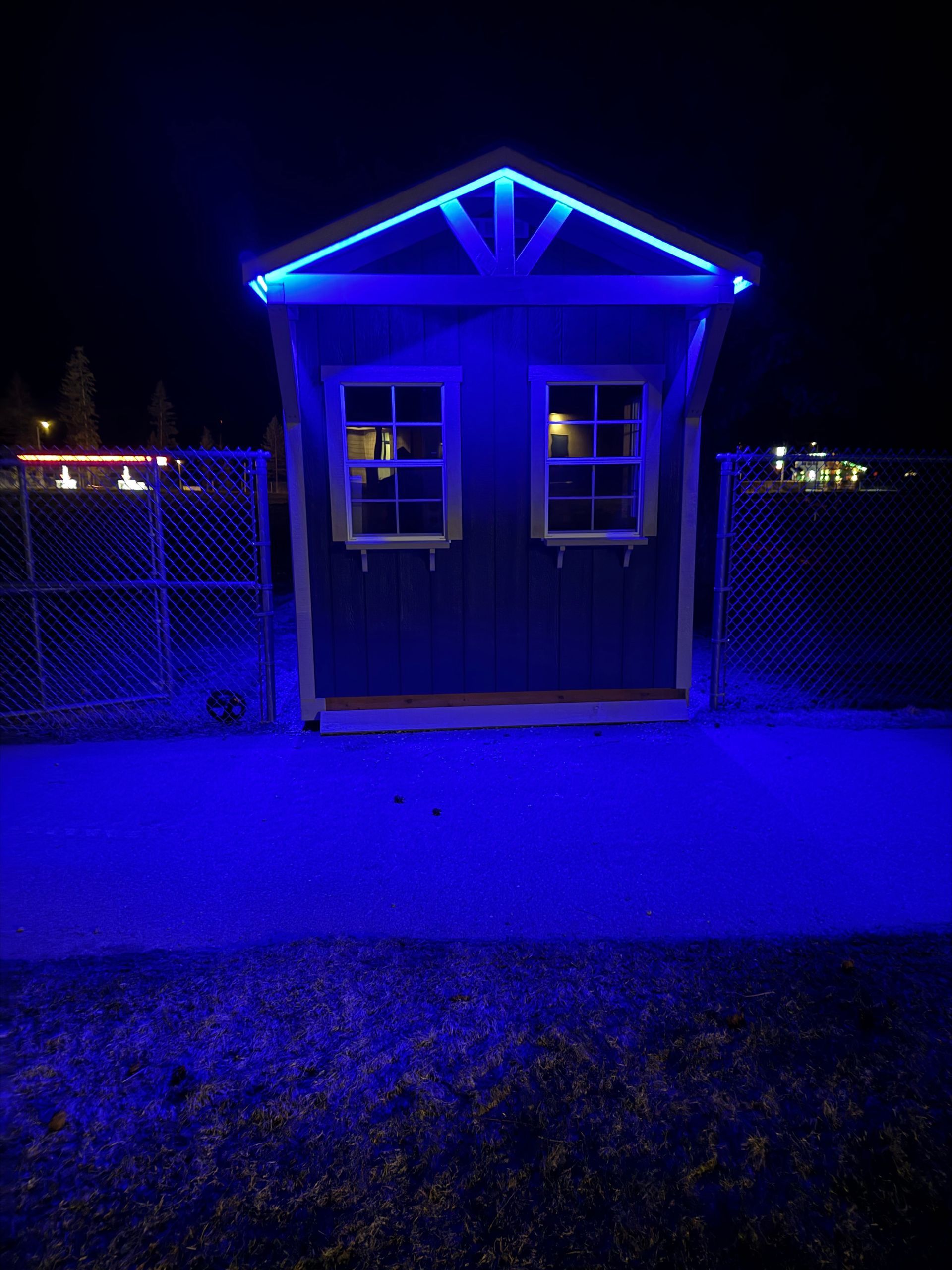 Blue-lit small shed or kiosk at night with two windows and a peaked roof, centered behind a fence.