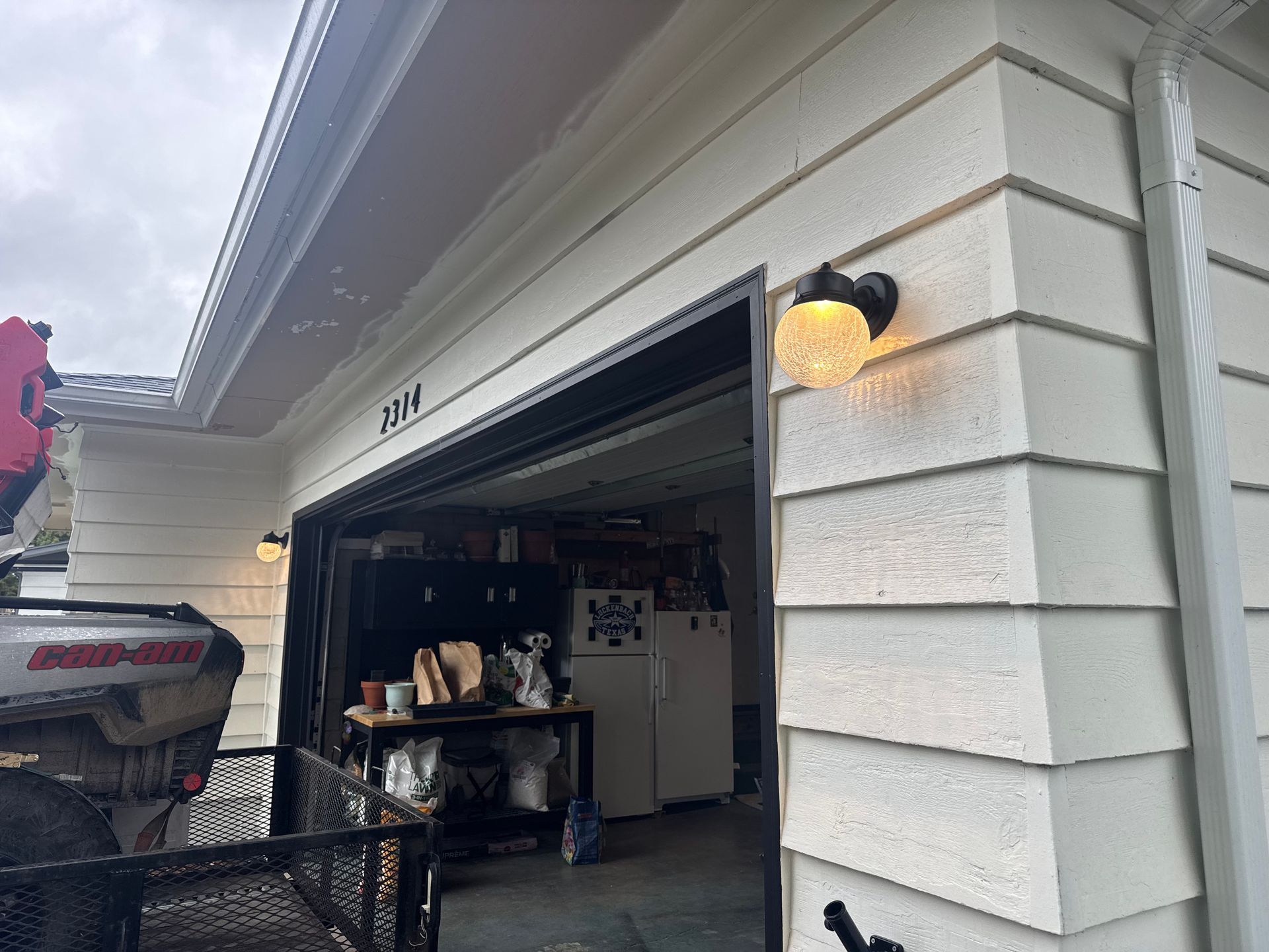 Open garage with workbench inside and a lit wall sconce on a white house exterior