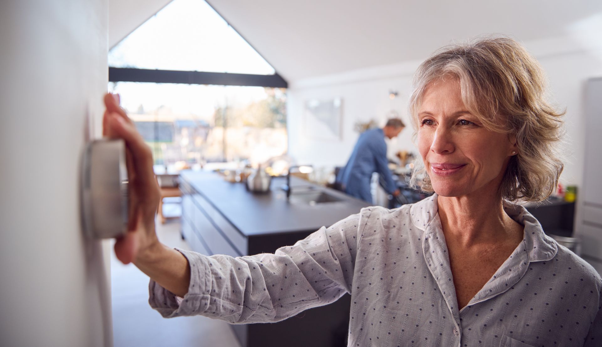 Woman adjusting a thermostat in a modern kitchen, smiling. A person is in the background.