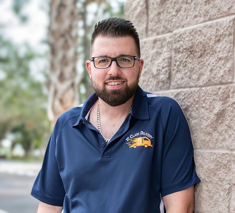 Man in blue polo shirt smiles, leans against a brick wall.