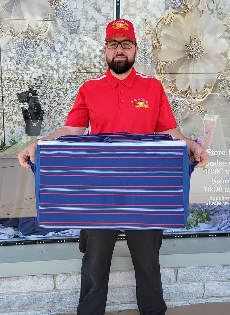 Man in red uniform holding a large blue and white striped package, outdoors.