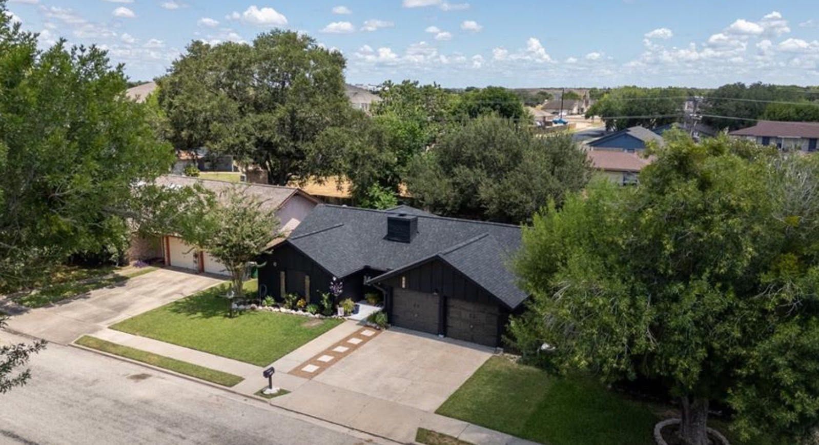 Suburban house with dark roof and front lawn, seen from above on a tree-lined street