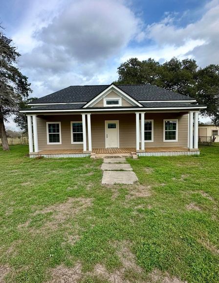 Single-story tan house with white porch columns, dark roof, and a front walkway on a grassy yard under cloudy sky