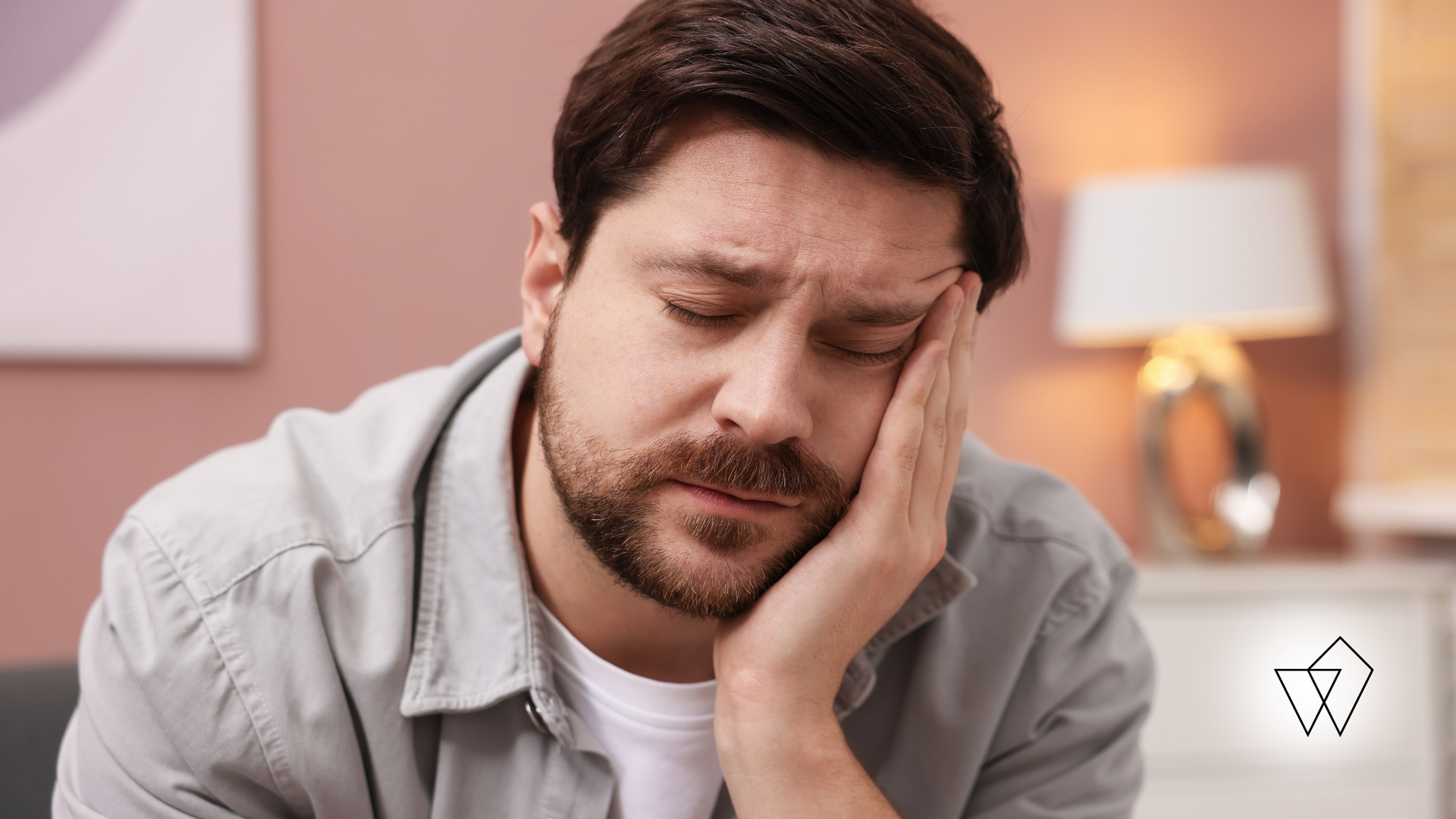 Man sitting on a bed, holding his face with a worried expression in a softly lit bedroom.
