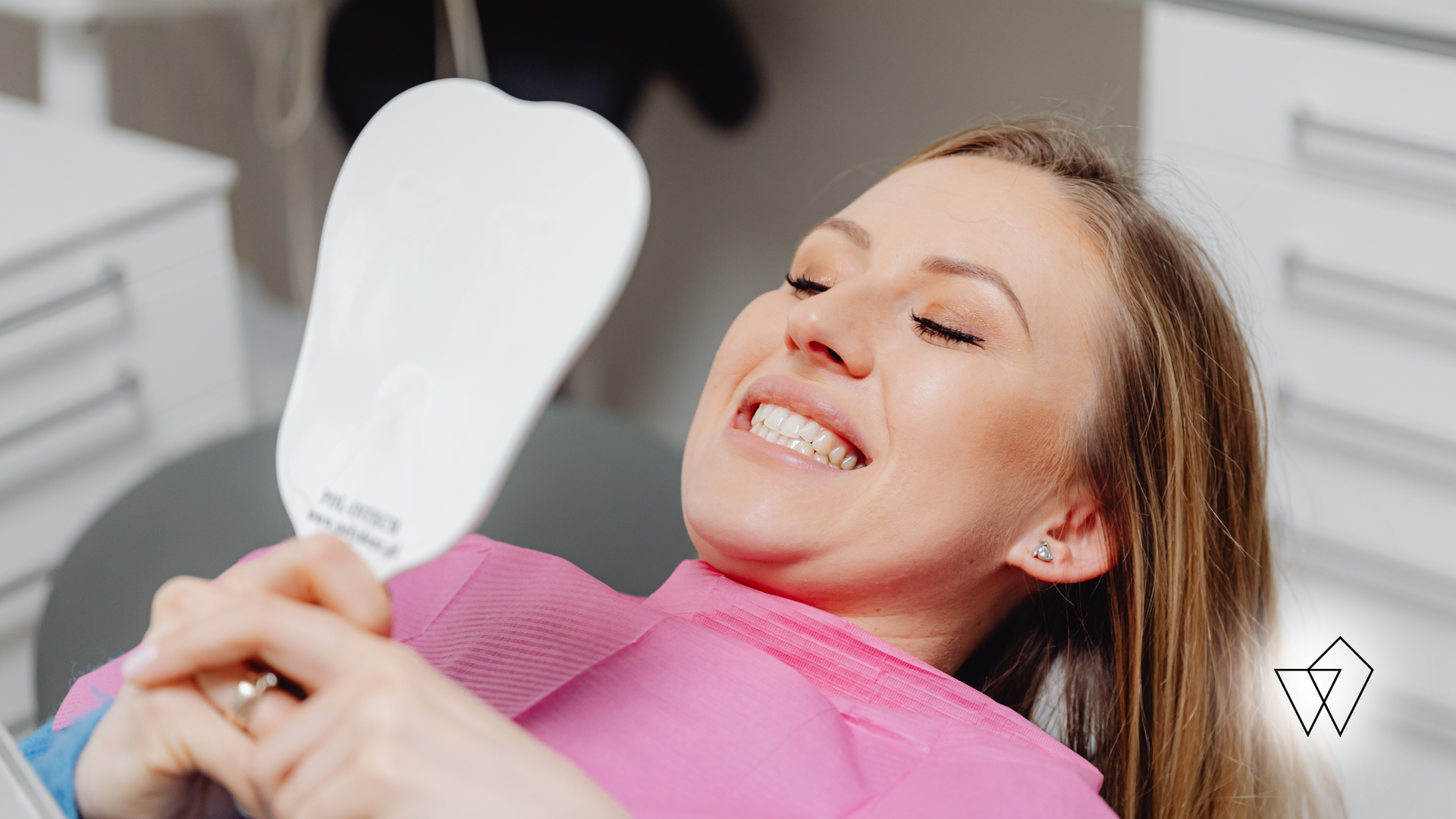 Woman smiling while holding a dental shade guide during a teeth whitening procedure