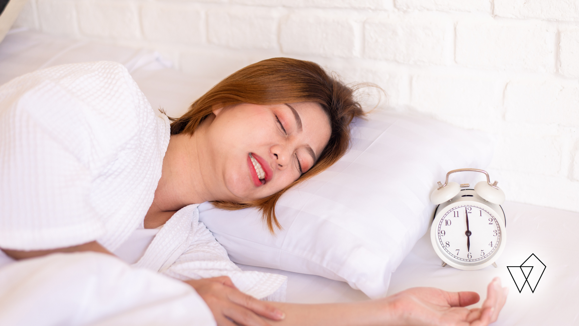 Sleeping person in white pajamas beside a white alarm clock on a bed