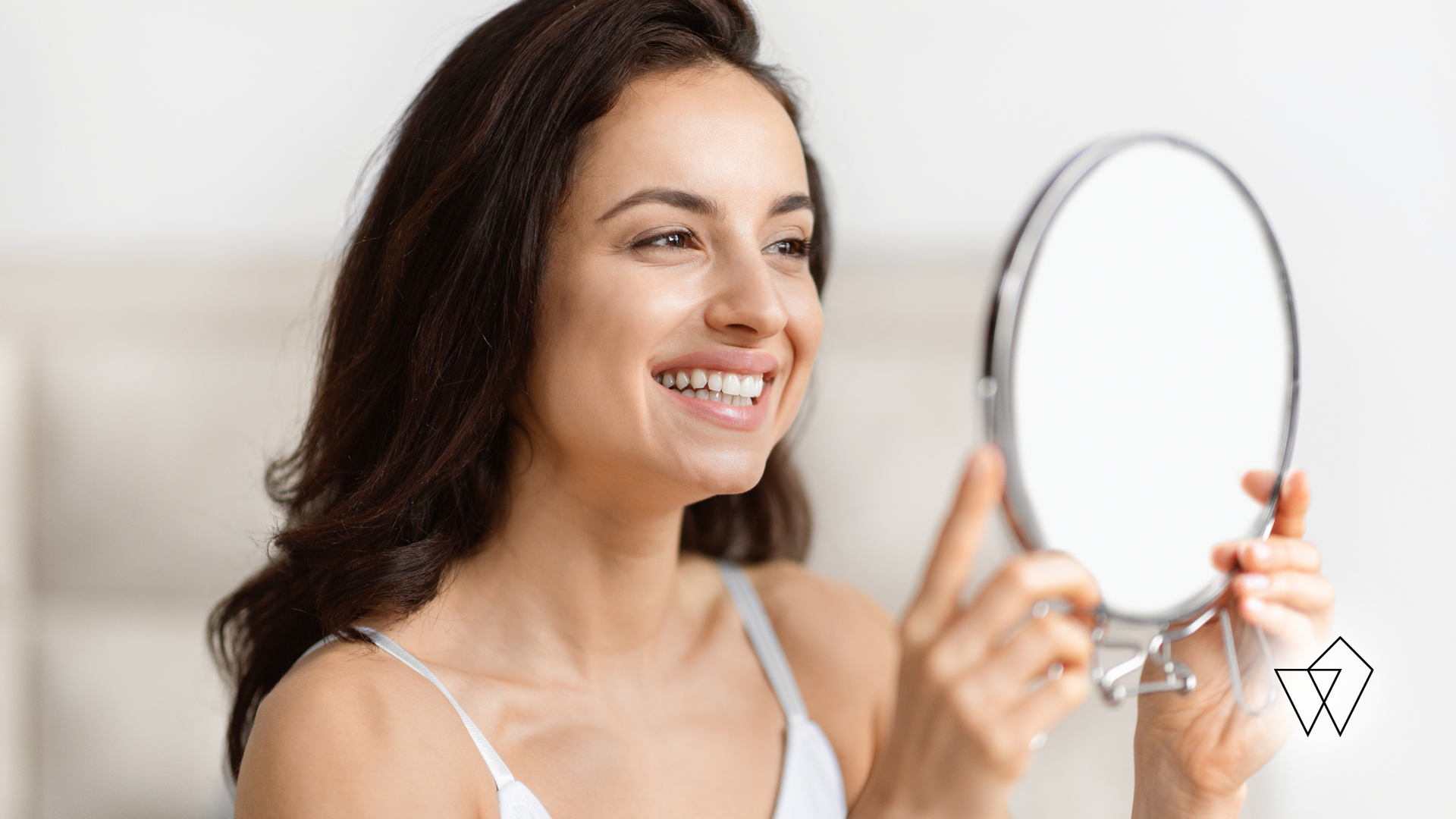 Smiling woman holds a round mirror while looking at her reflection in a bright room