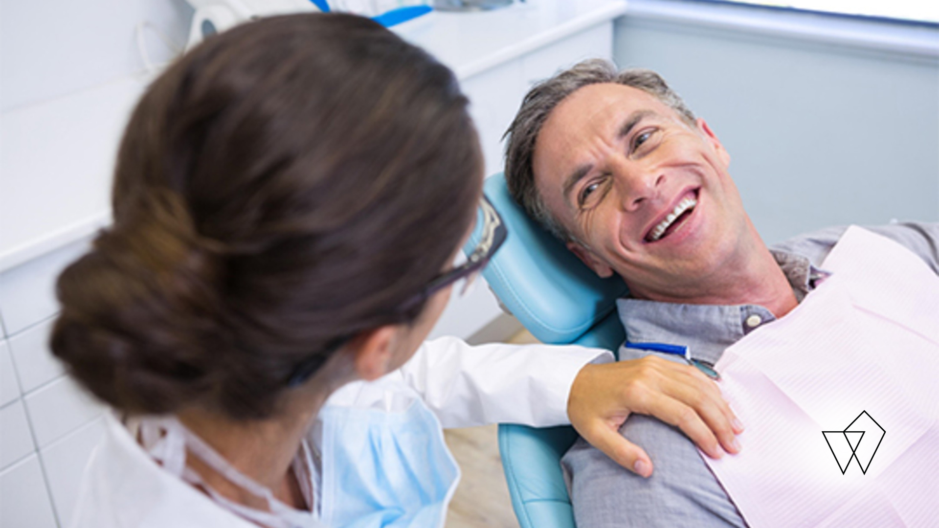 Patient reclining in a dental chair while a dentist examines him with a tool, both smiling.