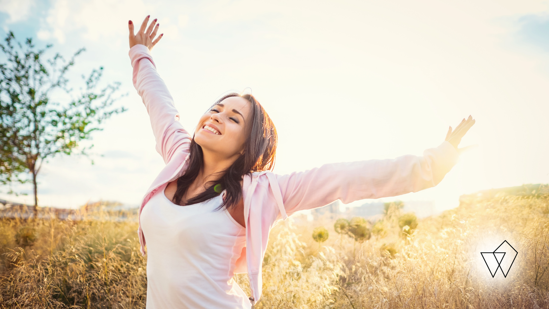 Woman in a pink jacket stands in a sunlit field with arms raised, smiling at the sky.