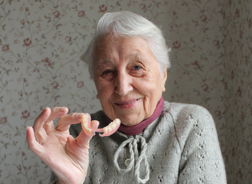 Elderly person holding dentures, smiling in front of floral wallpaper.