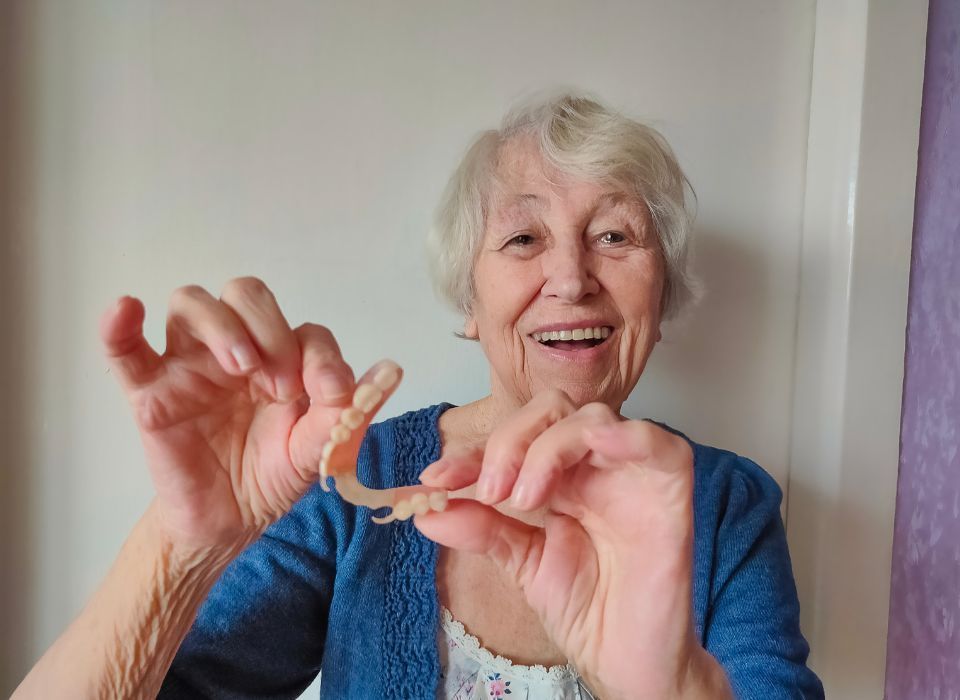 Smiling person holding partial dentures; indoors, neutral background.