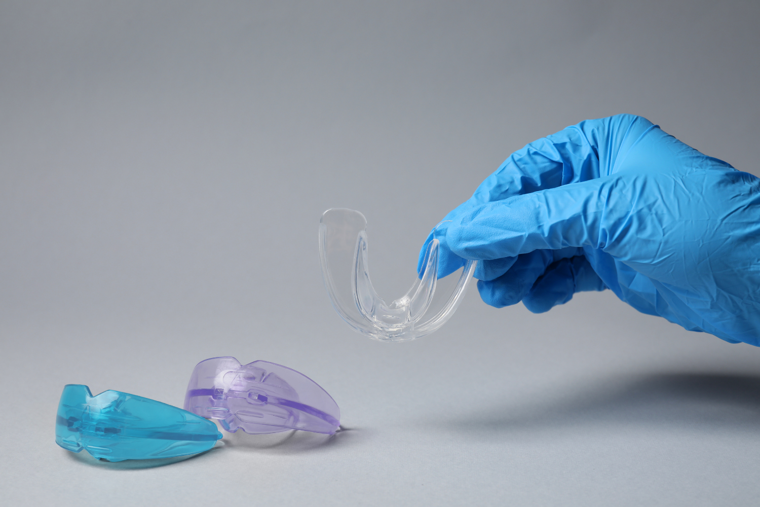 Gloved hand holding clear dental retainer above two colored molds on gray backdrop.
