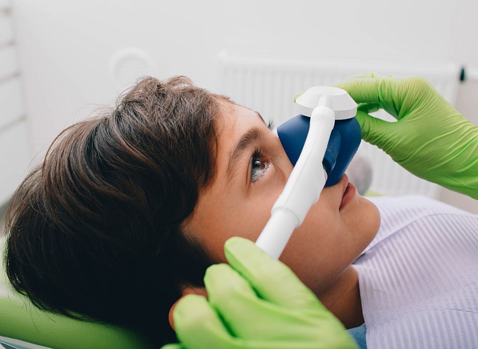 Dentist and assistant examining patient's teeth in a dental office. Both wear masks, patient reclined.