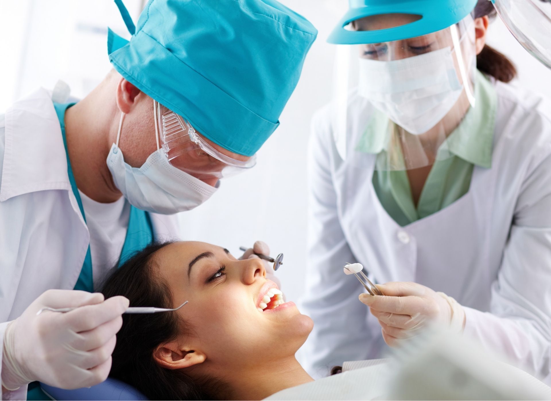 Dentist examining a patient's teeth. Assistant holding tools. All wearing masks, gloves, and scrubs.