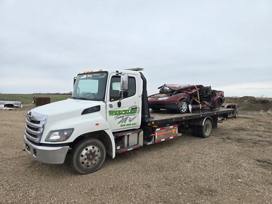 White tow truck carrying a damaged red car on a flatbed in a gravel lot under cloudy skies