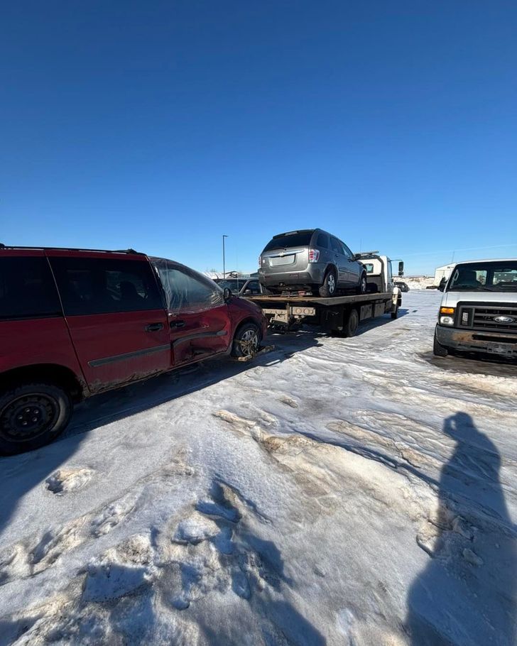 Snowy parking lot with a red SUV, a car on a tow truck, and a white van under a clear blue sky