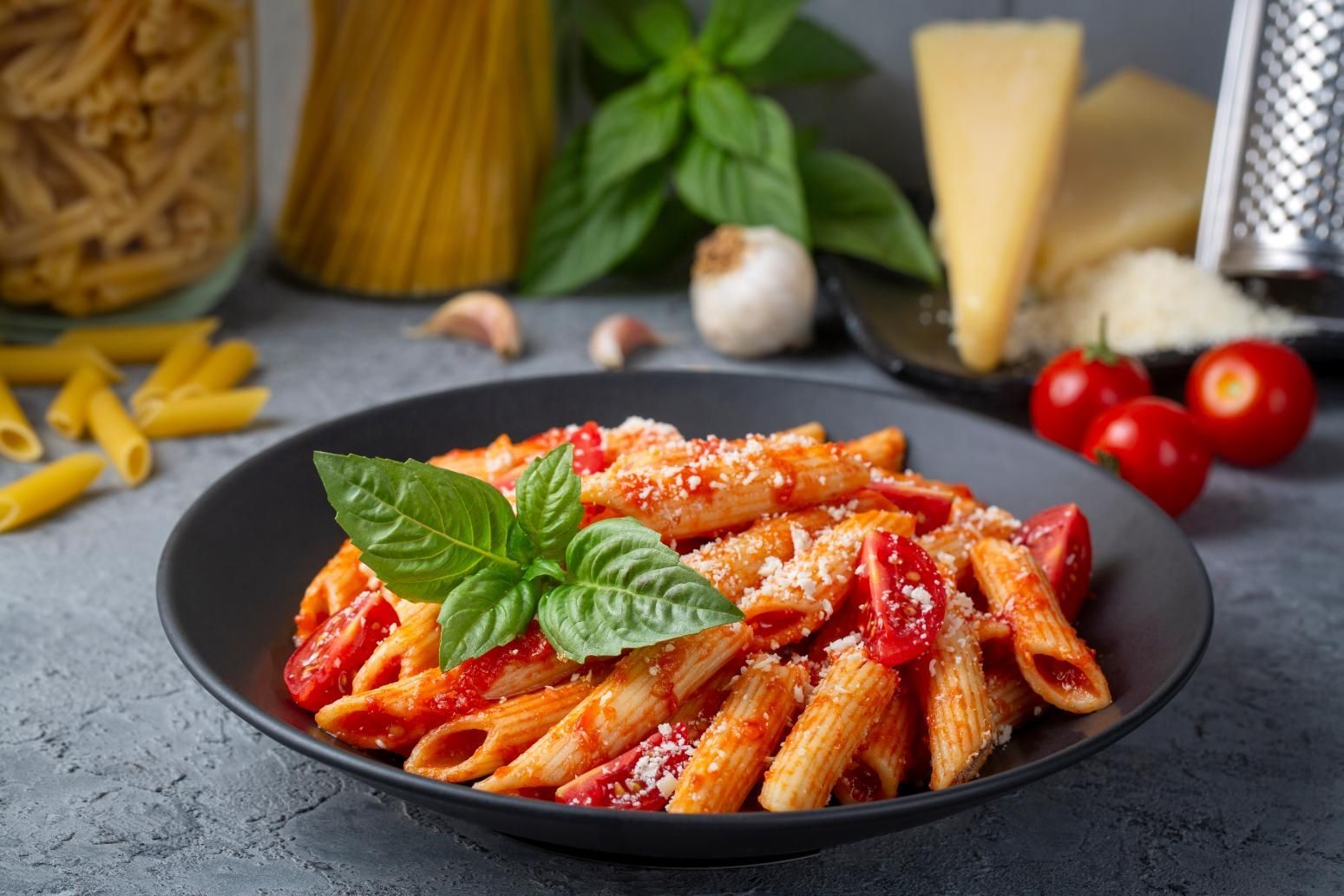 Penne pasta with tomato sauce, basil, and Parmesan cheese, in a black bowl, with ingredients in background.
