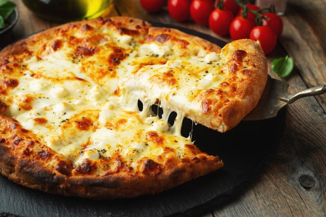 Pizza slice being lifted, cheese stretching, on a dark plate, tomatoes in background.