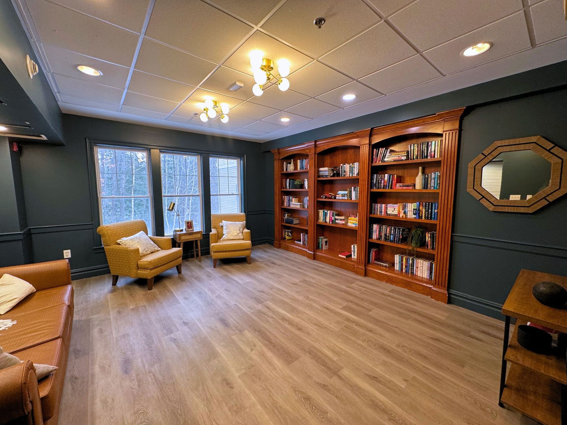 Cozy library with bookshelves, armchairs, and windows on a wood-look floor. Dark blue walls, a light ceiling, and a brown leather couch.