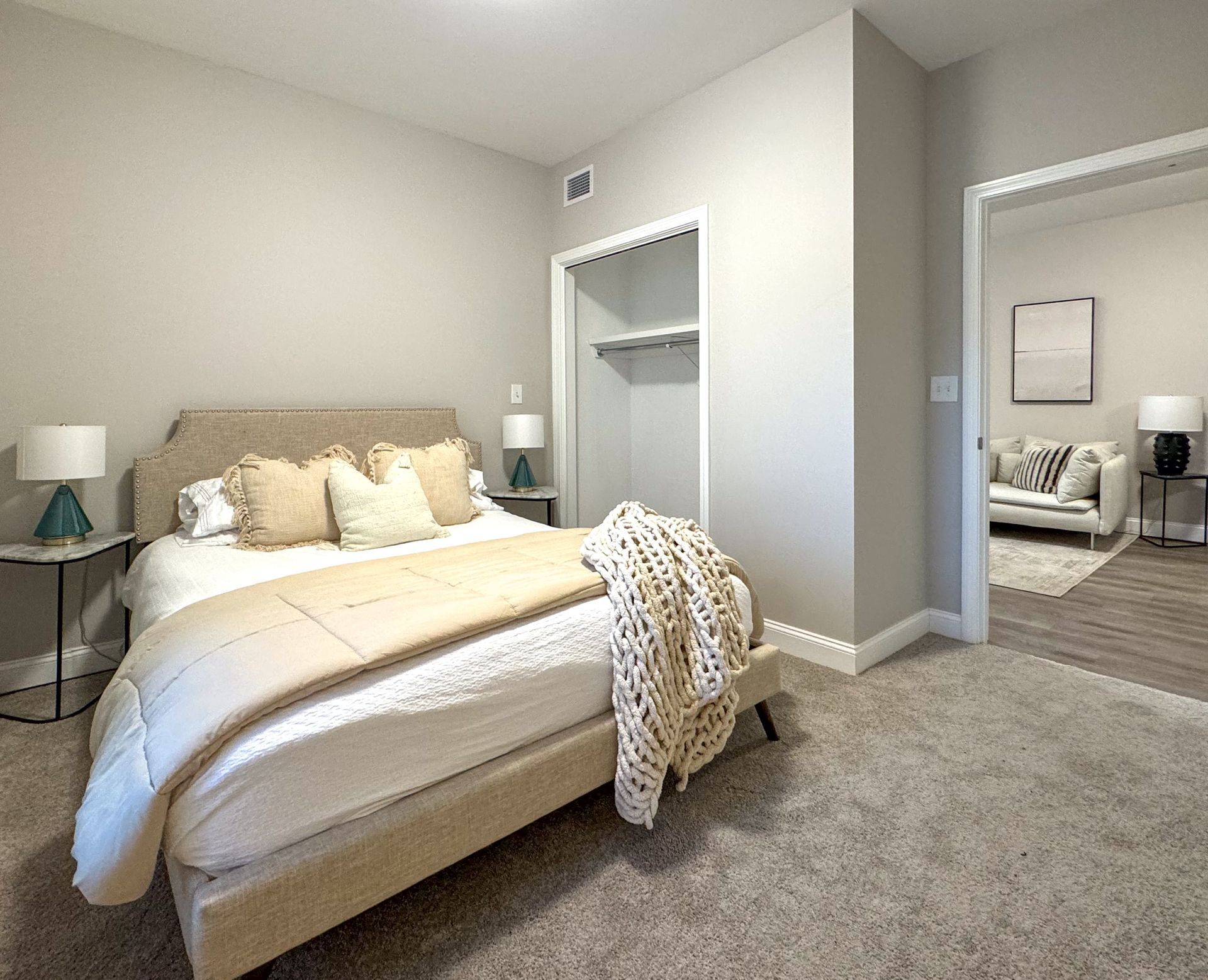 Bedroom with bed, nightstands, and open closet. Beige walls, gray carpet, and a view into a living room.